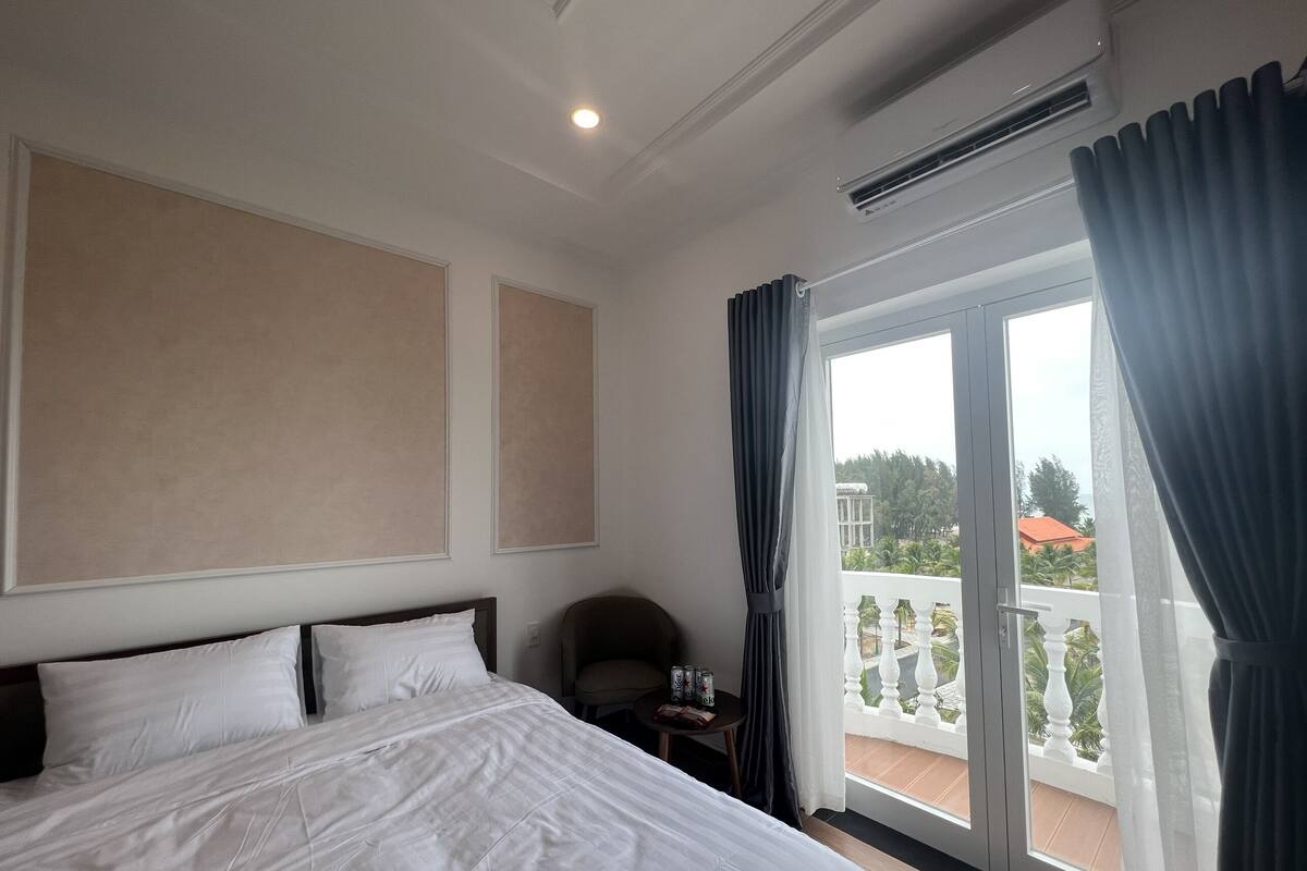 A hotel room with a large bed, a chair, a small table with drinks, and a balcony with a view of greenery and buildings.