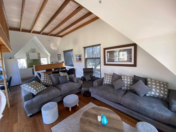 A spacious living area with a high ceiling and wooden beams, featuring a grey sectional sofa, a coffee table, and a kitchen in the background.