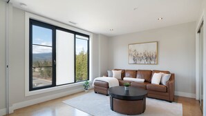 A spacious living room with a large window, a brown leather sofa, a round coffee table, and a painting on the wall. - Luxury Condo on Mara Lake with Boat Slip & Pool. (Sicamous)