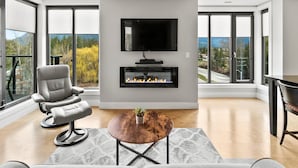 A modern living room with a fireplace, a comfortable seating area, and large windows offering a scenic view. - Luxury Condo on Mara Lake with Boat Slip & Pool. (Sicamous)
