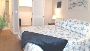A bedroom with a bed, a nightstand, a lamp, a door, and a wall with a framed picture.