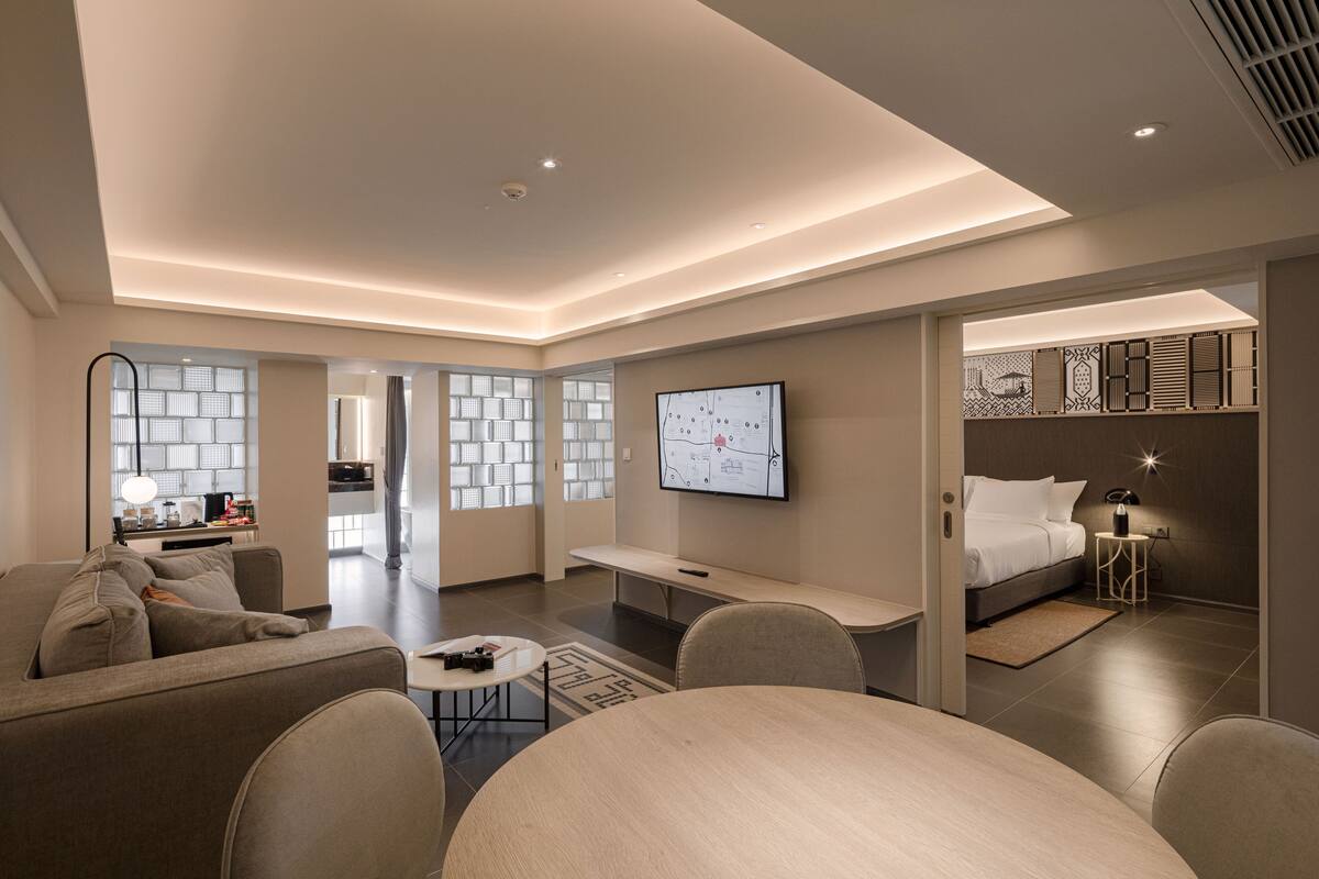 A modern hotel room with a sofa, a small table, a TV mounted on the wall, and a bed with a nightstand.