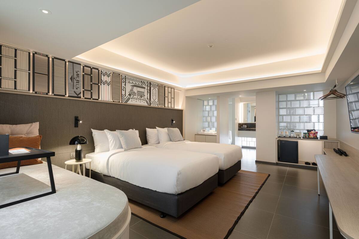 A modern hotel room with two beds, a desk, a small kitchen area, and a bathroom.