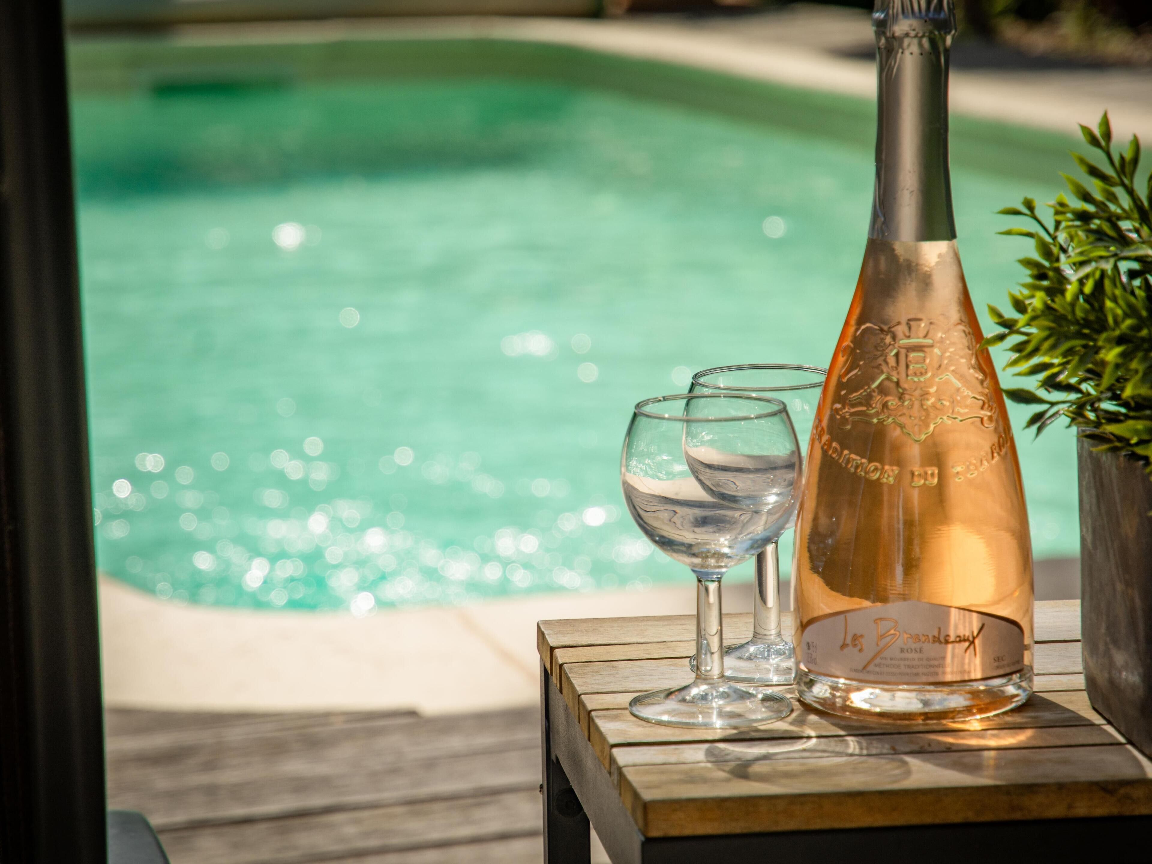 A bottle of champagne and two glasses on a wooden table by a pool.