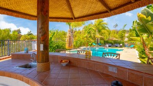 A poolside area with a tiled deck, a thatched umbrella, and a view of a swimming pool surrounded by greenery.