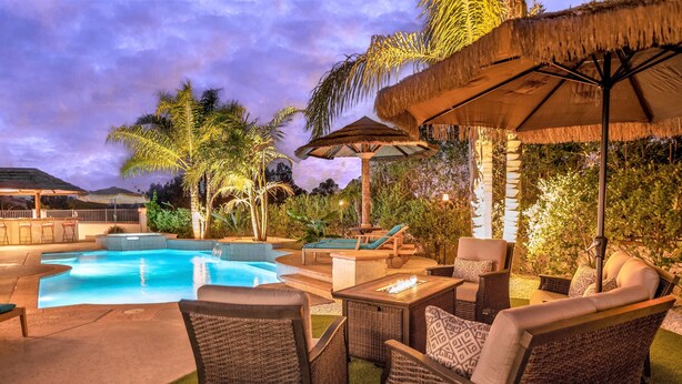 A swimming pool with lounge chairs and a fire pit, surrounded by tropical plants and thatched umbrellas.