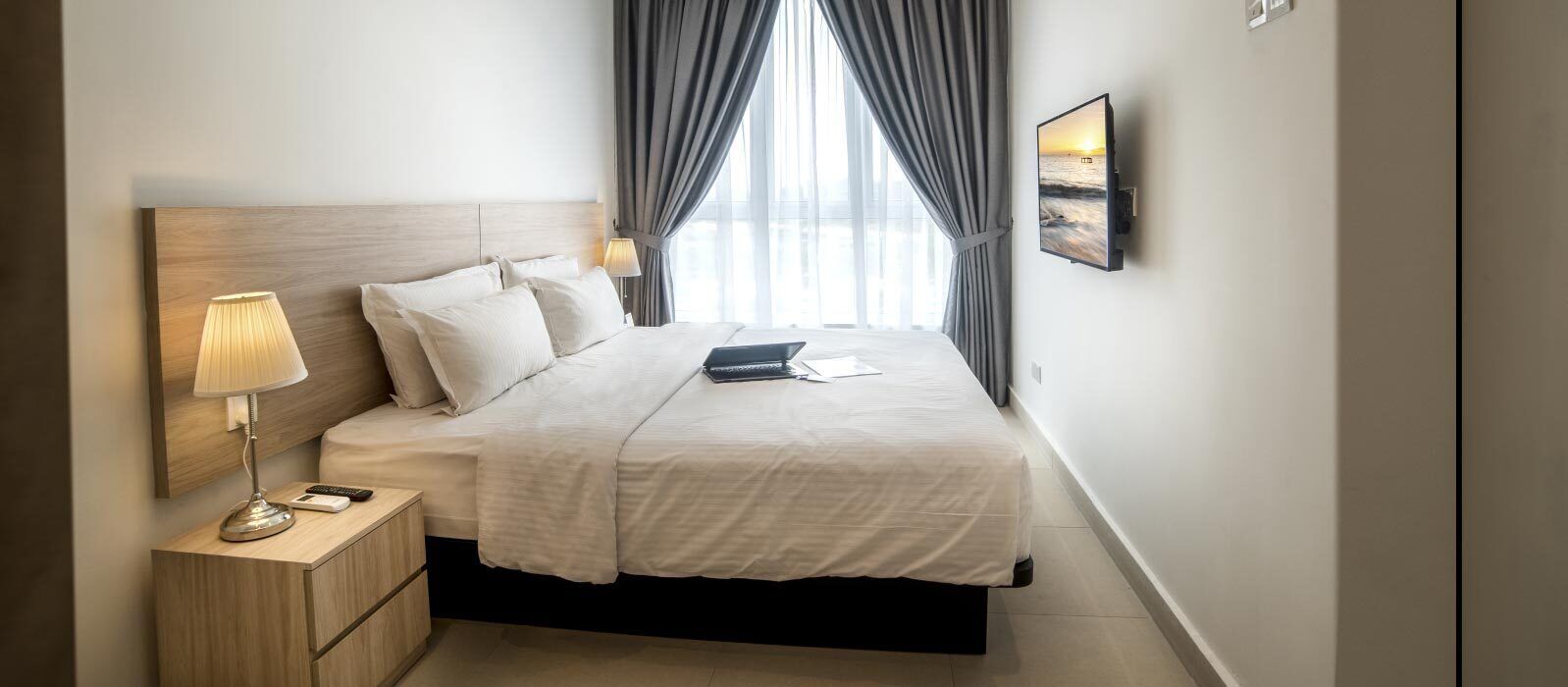 A hotel room with a bed, bedside table, lamp, and a TV on the wall.