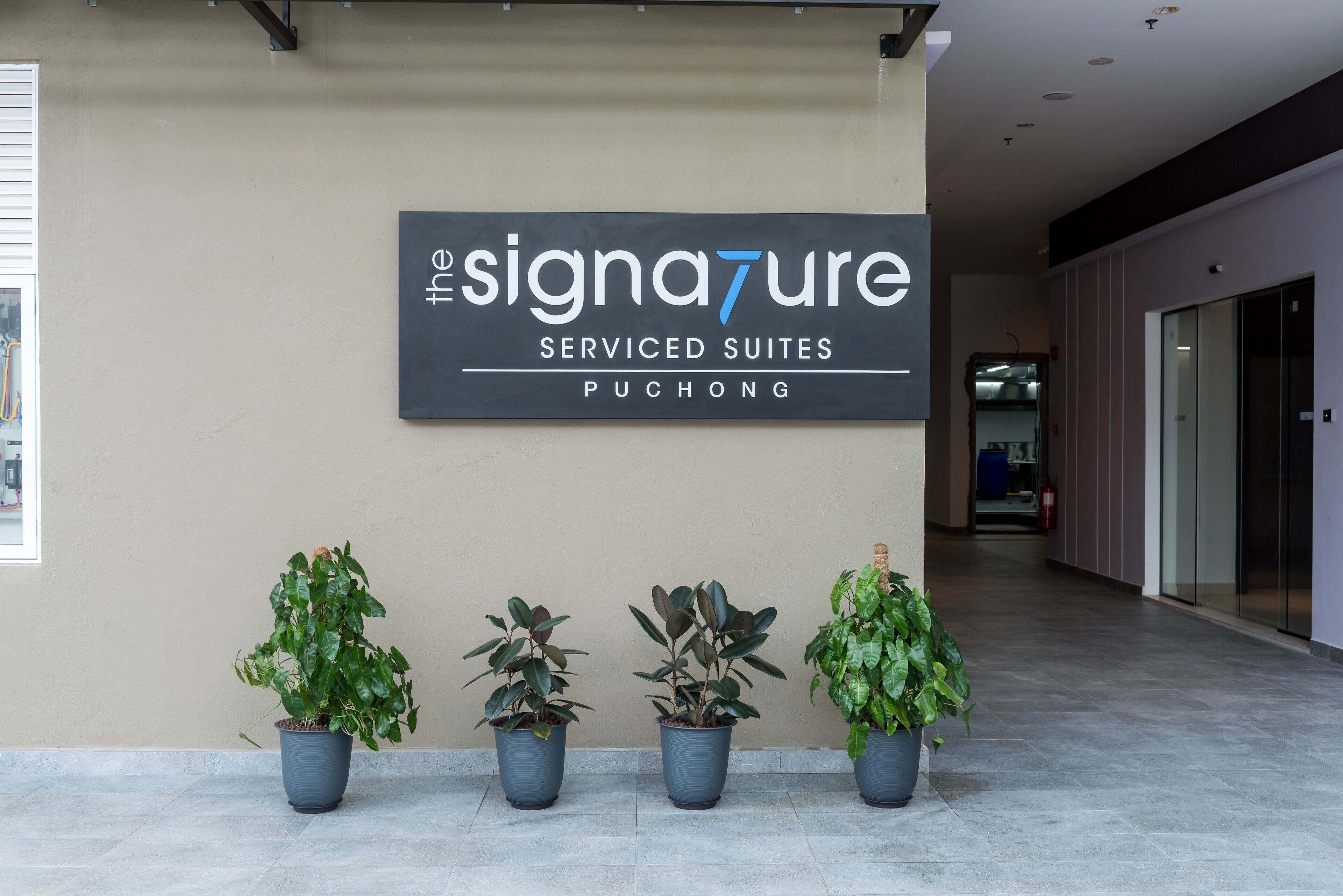 A reception area with a sign for 'The Signature Serviced Suites' in Puchong. There are three potted plants in front of the sign.