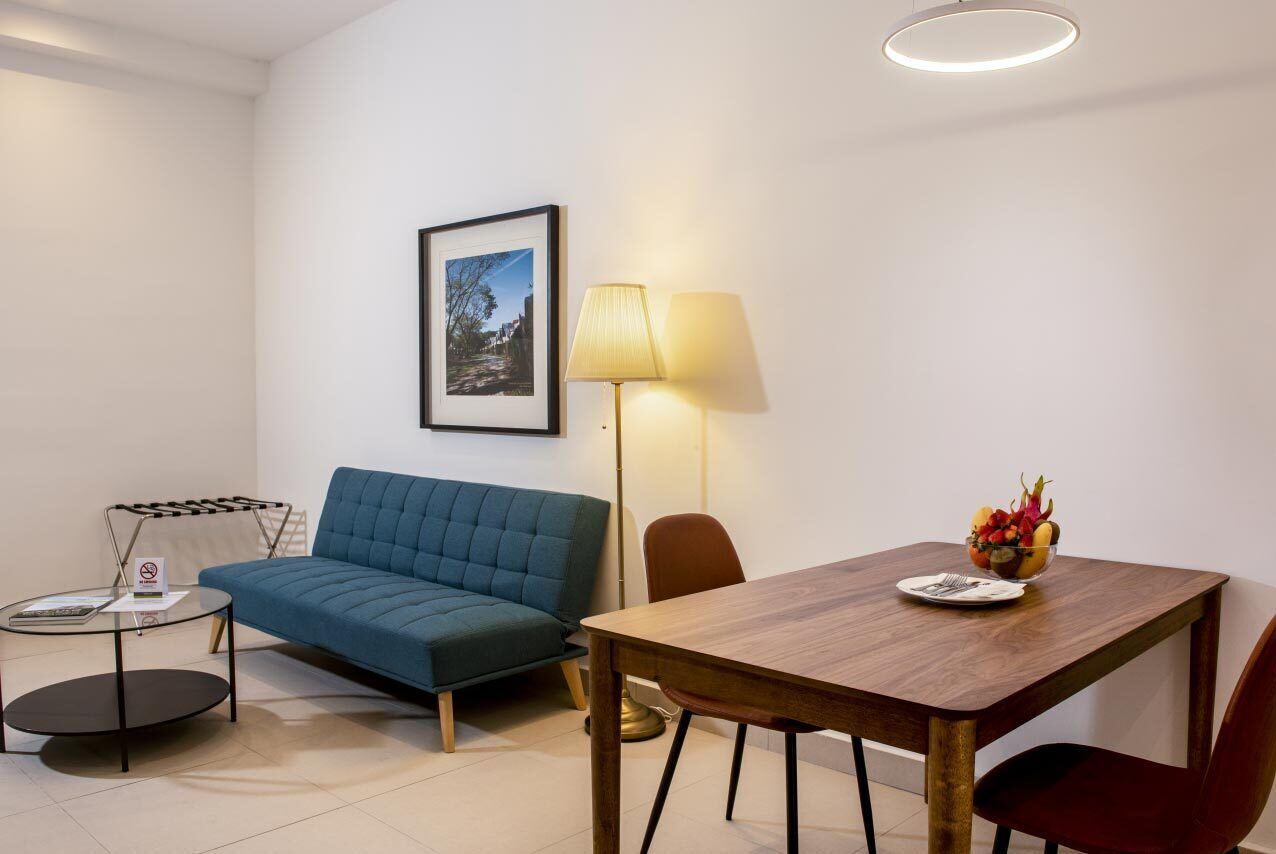 Un salon moderne avec un canapé bleu, une table à manger en bois sur laquelle se trouve un panier de fruits, un lampadaire et un tableau encadré accroché au mur.