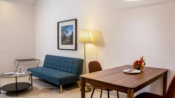 Un salon moderne avec un canapé bleu, une table à manger en bois sur laquelle se trouve un panier de fruits, un lampadaire et un tableau encadré accroché au mur.