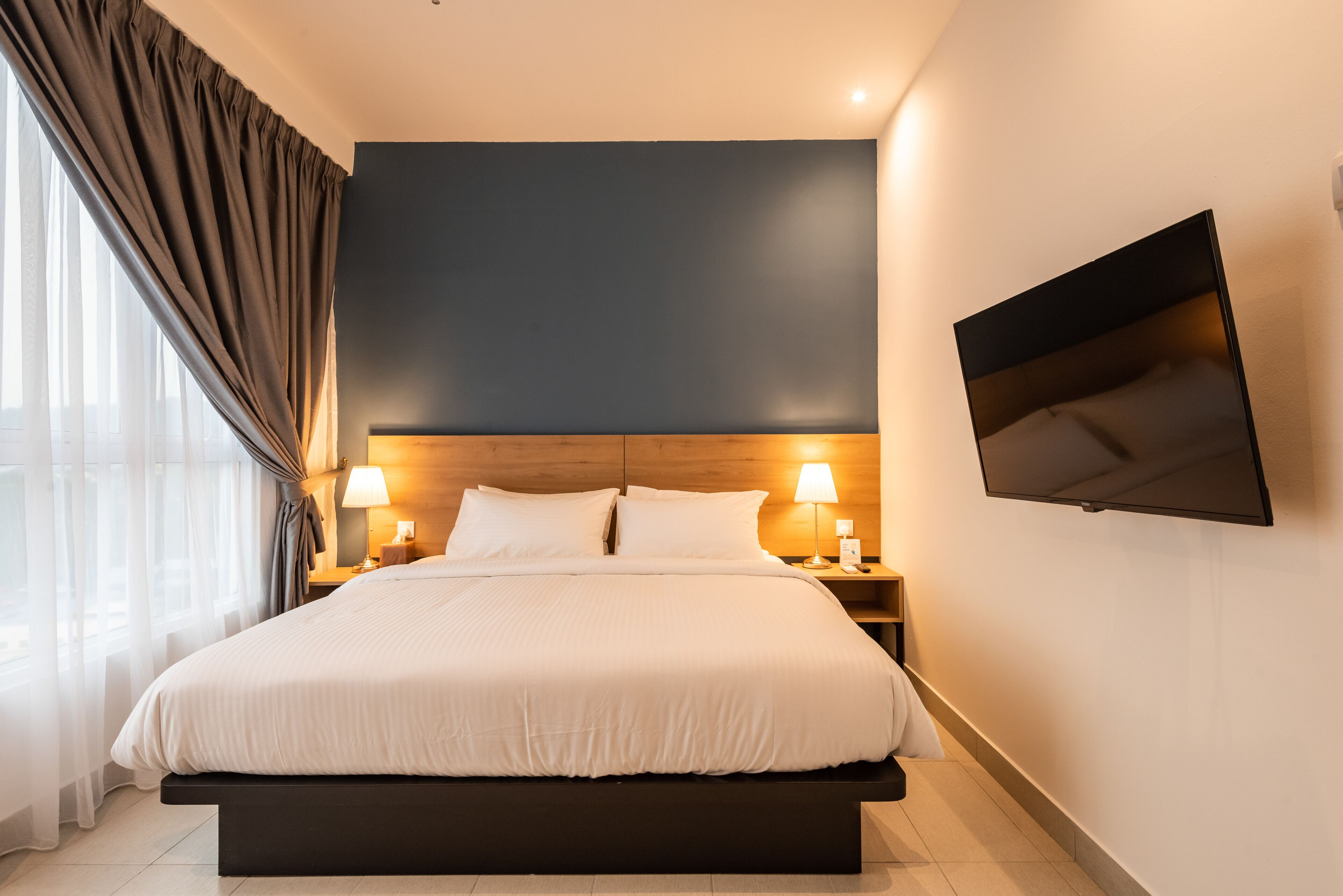 A modern hotel room with a large bed, bedside lamps, a flat-screen TV, and a view of the city through sheer curtains.