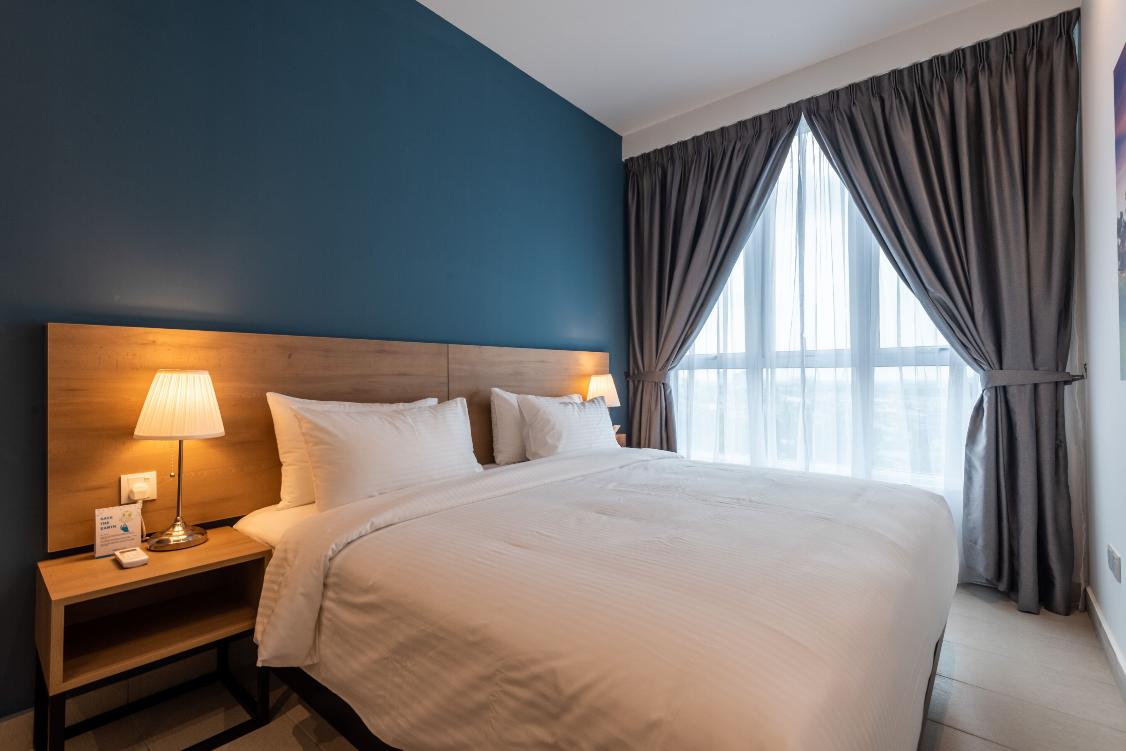 A hotel room with a large bed, a wooden headboard, a nightstand with a lamp, and a window with curtains.