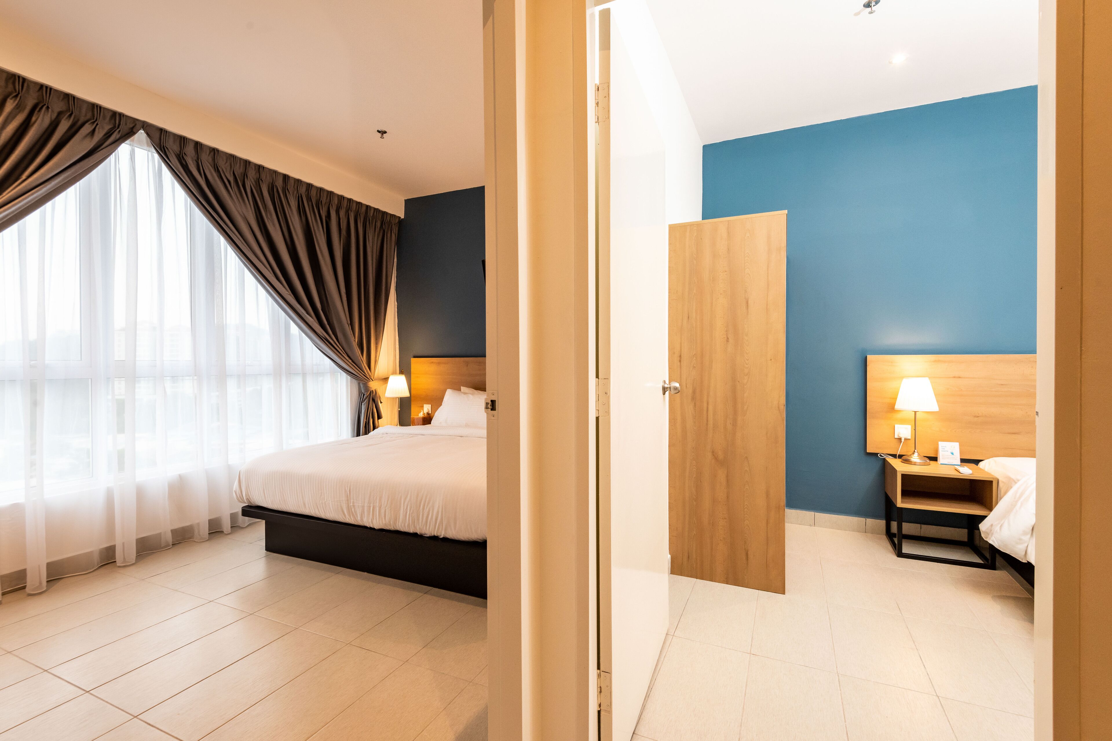 A modern hotel room with a large bed, a bedside table with a lamp, and a view through a window with sheer curtains.