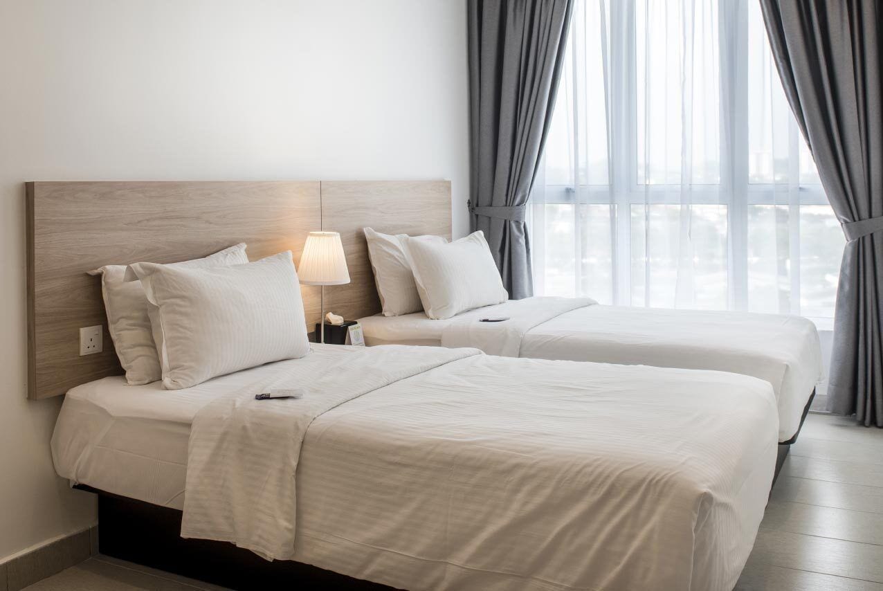 A hotel room with two beds, a wooden headboard, a bedside lamp, and a view of the city through large windows.
