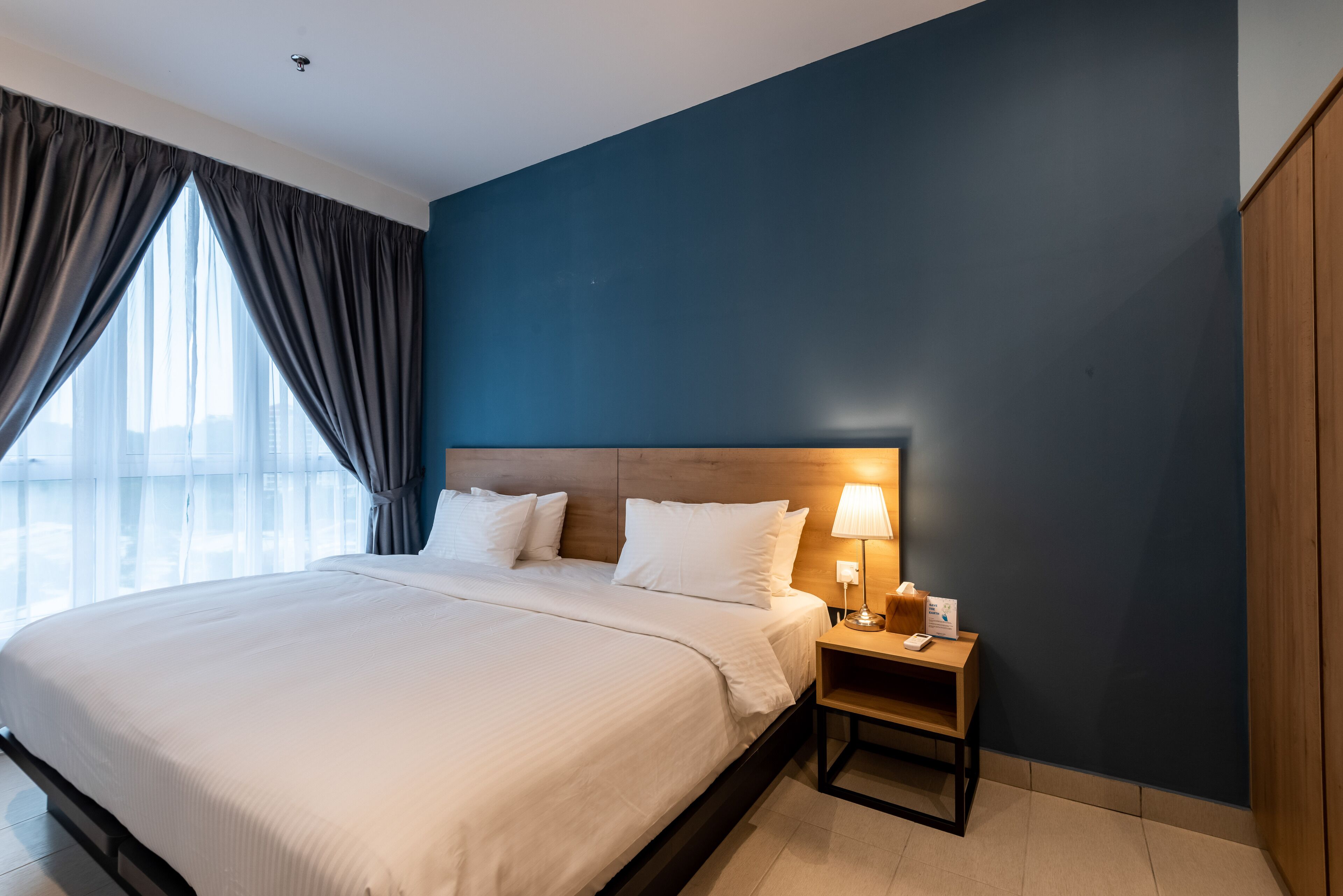 A hotel room with a large bed, a nightstand with a lamp, and a view of the city through the window.
