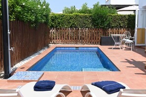 En rektangulär swimmingpool med en kant av kakel, omgiven av ett trästaket och en kakelbelagd uteplats med två solstolar och ett bord.