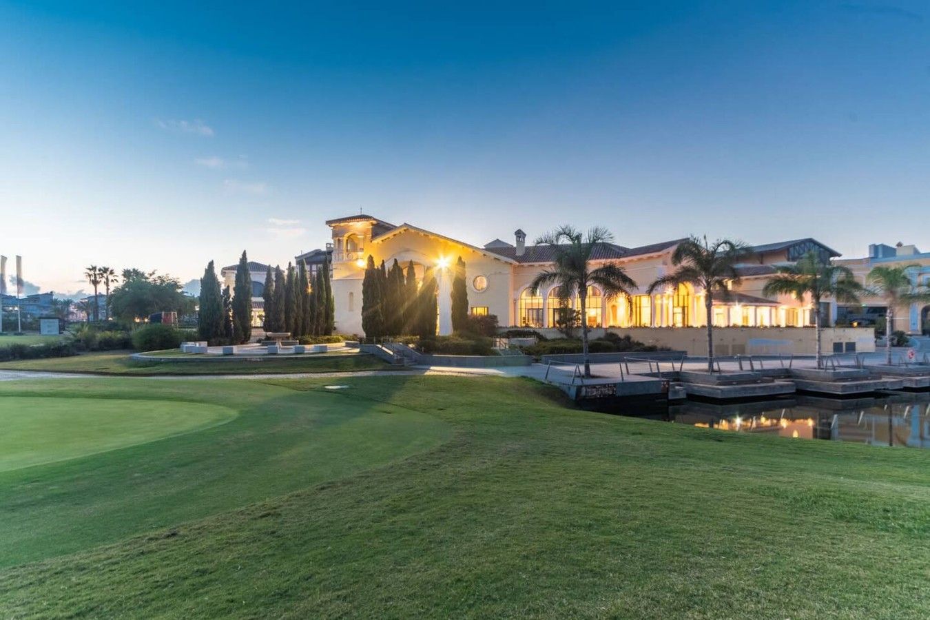 Un campo de golf bien cuidado con club house, palmeras y un estanque tranquilo.