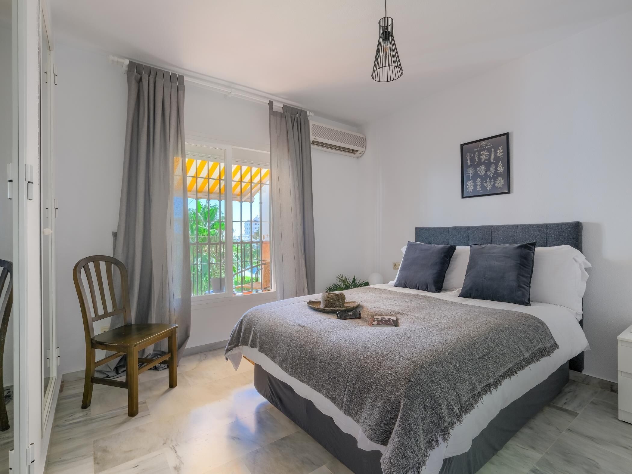 A bedroom with a bed, a chair, a window with curtains, an air conditioning unit, and a hanging light fixture.
