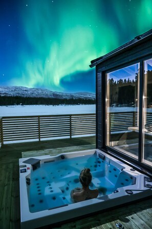A hot tub on a wooden deck with a view of a snowy landscape and the Northern Lights.