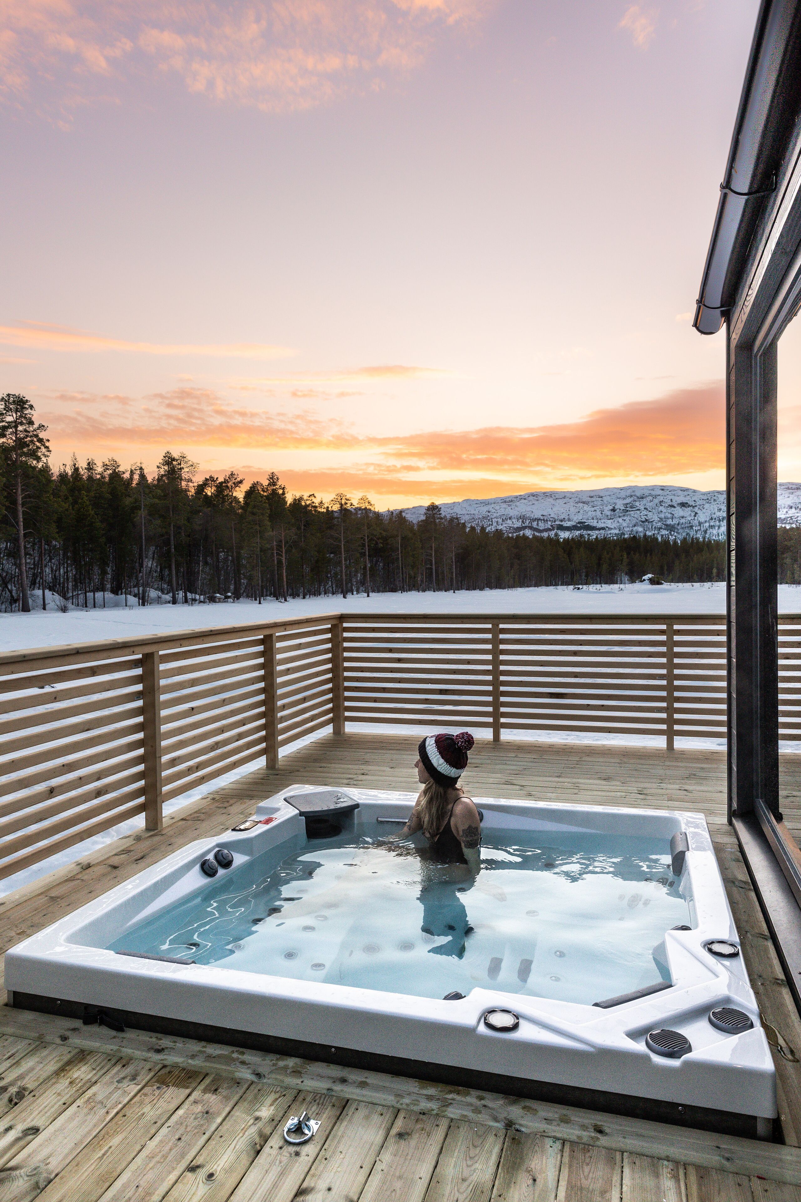Eine Holzterrasse mit Whirlpool mit Blick auf eine verschneite Landschaft und einen Sonnenuntergang.