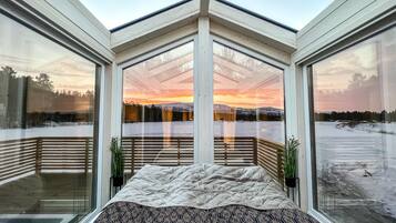 A modern bedroom with a large glass window offering a view of a snowy landscape and a sunset.