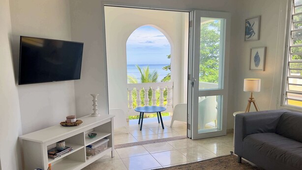 A living room with a TV, a sofa, a coffee table, and a view of the ocean through an open door. - Aguada Ocean Club 3A - 2 Bed 1 Bath Suite (Aguada)