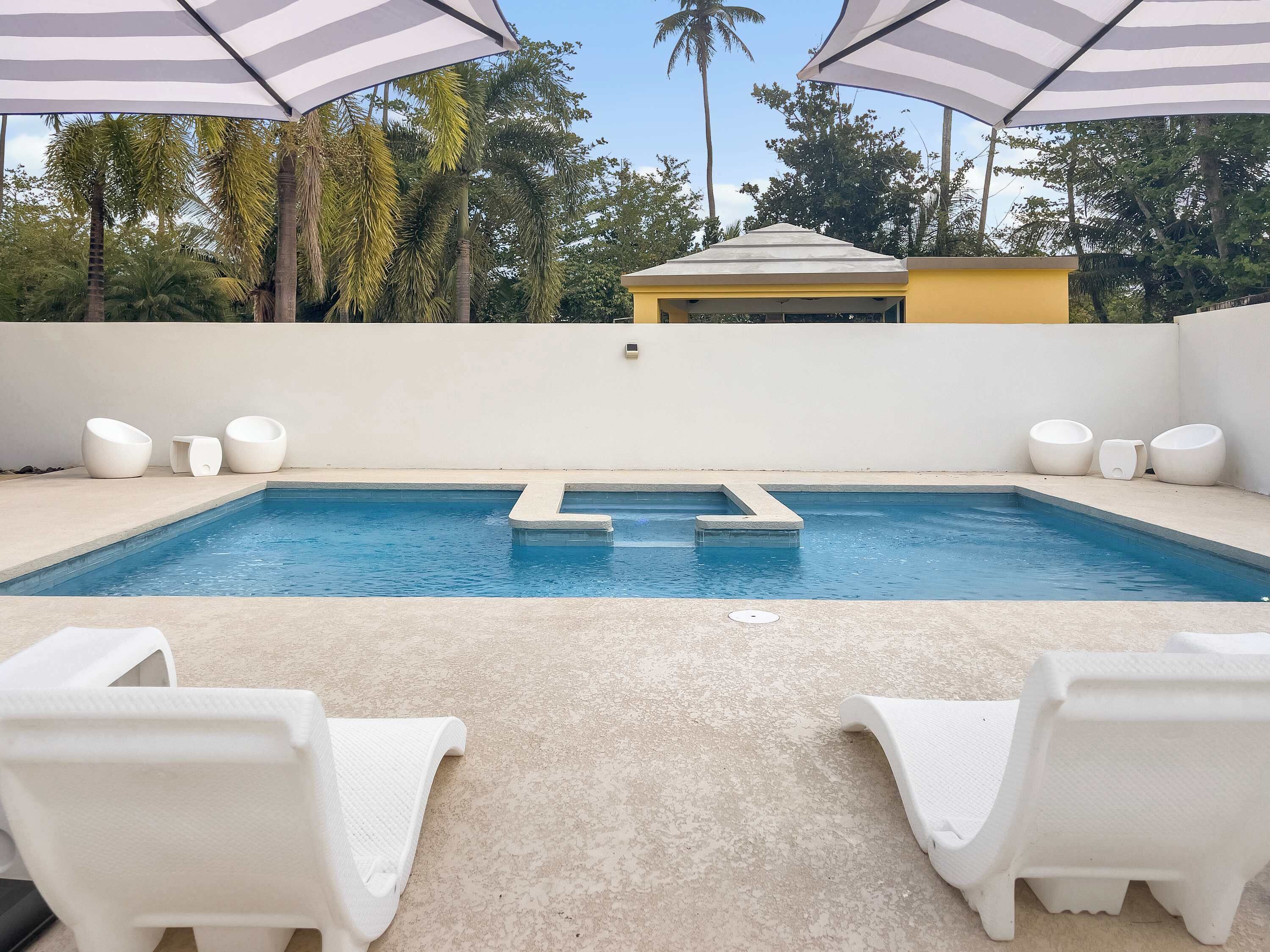 Una piscina con sillas de descanso blancas y sombrillas, rodeada de palmeras y un edificio amarillo.