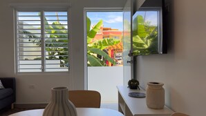 Una sala de estar moderna con un gran ventanal, un televisor de pantalla plana montado en la pared, una mesa de comedor y una mesita auxiliar con objetos decorativos.
