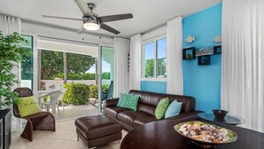 A modern living room with a brown leather sofa, a dining table with a decorative bowl, and a ceiling fan. There is a glass door leading to a balcony with outdoor furniture. - Blue Water View | Breathtaking 2 bedroom apartment with amazing water views! (Rincon)