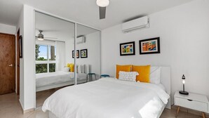 A modern bedroom with a large bed, a ceiling fan, a bedside table with a lamp, and a window with curtains. - Blue Water View | Breathtaking 2 bedroom apartment with amazing water views! (Rincon)
