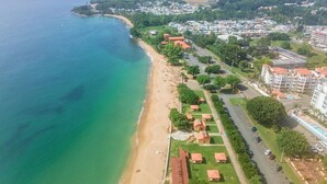 An aerial view of a coastal area with a beach, a row of buildings with red roofs, and a road running parallel to the beach. - Blue Water View | Breathtaking 2 bedroom apartment with amazing water views! (Rincon)
