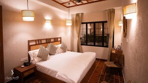 A hotel room with a large bed, two bedside lamps, a wooden headboard, a nightstand, a chair, a window with curtains, and a ceiling fan. - Shanti Inn Dormitory (Varanasi)