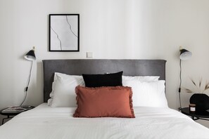 Uma cama bem arrumada com almofadas brancas e pretas, uma almofada de destaque marrom e uma obra de arte abstrata emoldurada na parede.