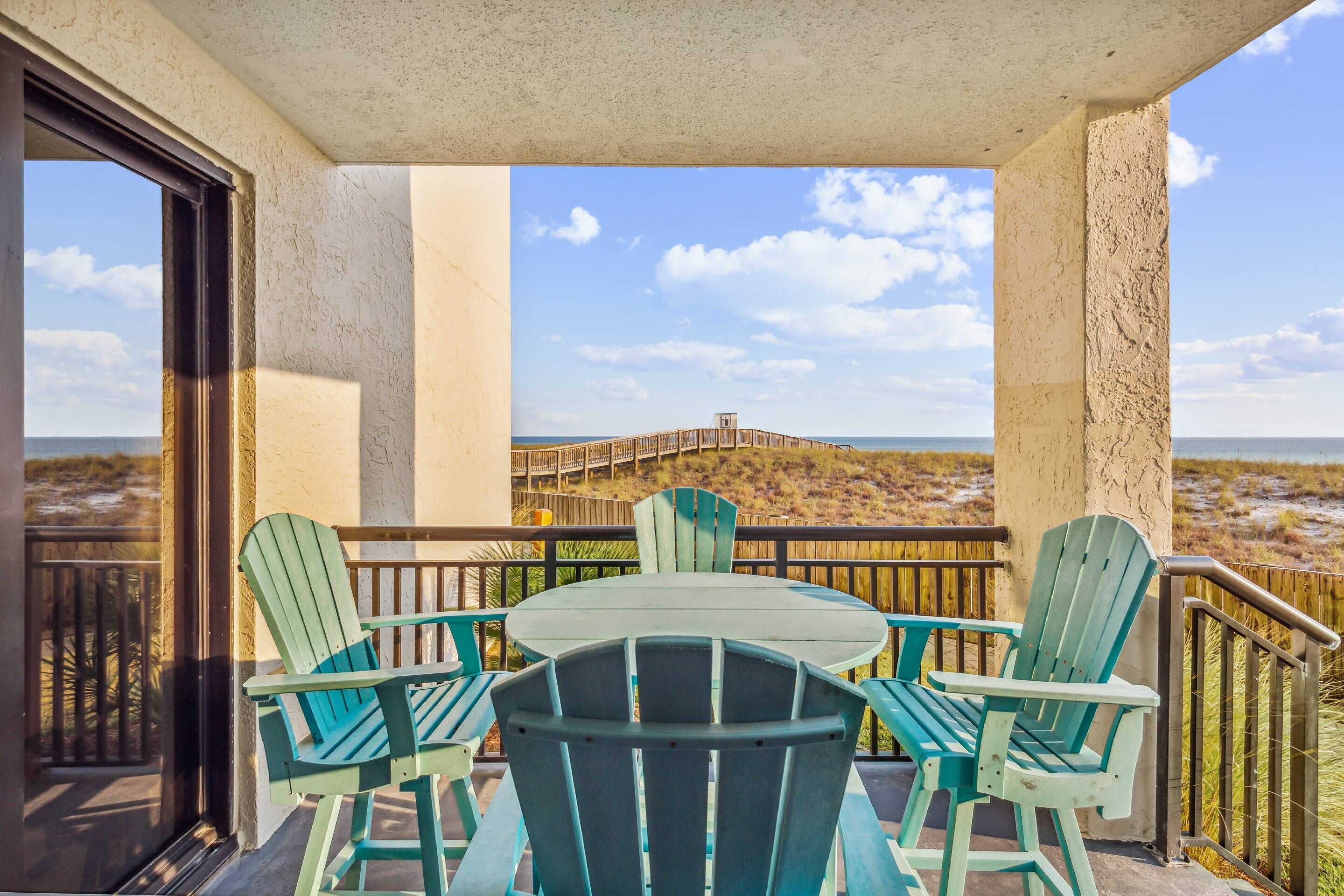 A balcony with a table and chairs, overlooking a beach and a boardwalk.