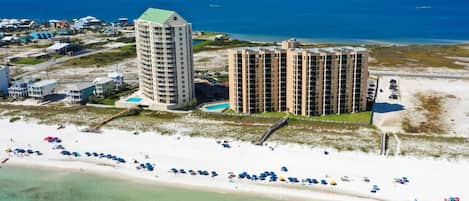An aerial view of a coastal area with two large buildings, a beach, and a clear blue sea.