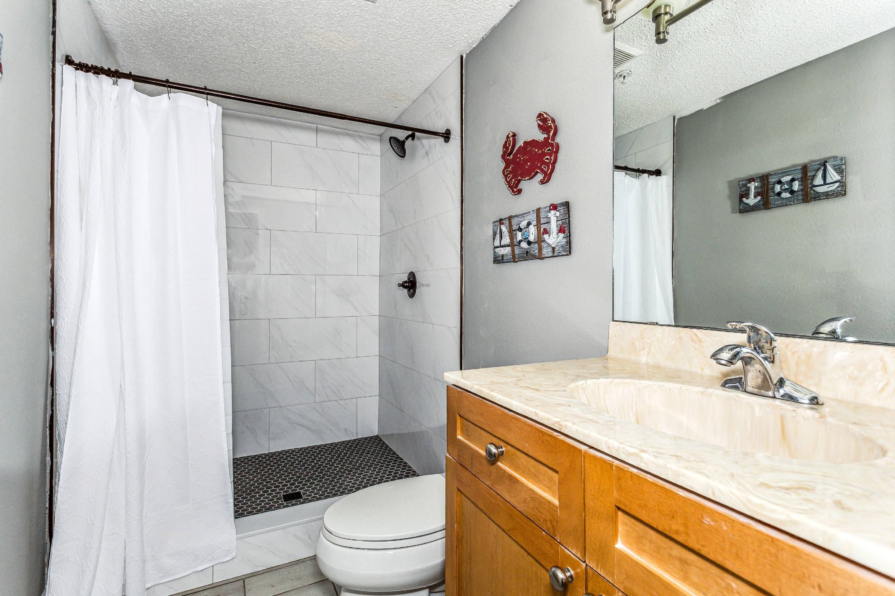 Une salle de bain avec un rideau de douche blanc, des toilettes blanches, un meuble-lavabo en bois avec un plan de travail en marbre et un miroir fixé au mur.