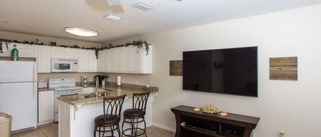A kitchen with white cabinets, a microwave, stove, and a ceiling fan. There is a bar area with stools and a TV mounted on the wall.