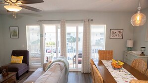 Un salon comprenant un canapé, un fauteuil et une table à manger avec des chaises en osier. On y trouve également un ventilateur de plafond, une fenêtre avec des stores et une porte donnant sur l’extérieur.