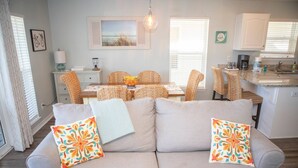 Un salon avec un canapé, des coussins colorés, un coin repas avec des chaises en osier, une cuisine avec des meubles blancs et un luminaire suspendu.