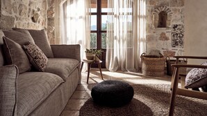 Una sala de estar acogedora con una pared de piedra, un ventanal de madera, un sofá, un otoman redondo y un sillón de mimbre.