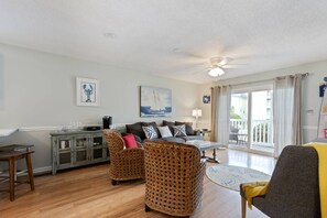 A living room with a sofa, two wicker chairs, a coffee table, and a dining table. There is a ceiling fan, a painting of a sailboat, and a view of a balcony with a railing.