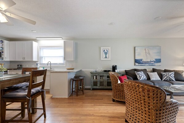A modern kitchen with white cabinets, a dining area with a wooden table and chairs, and a living room with a sofa and wicker chairs.