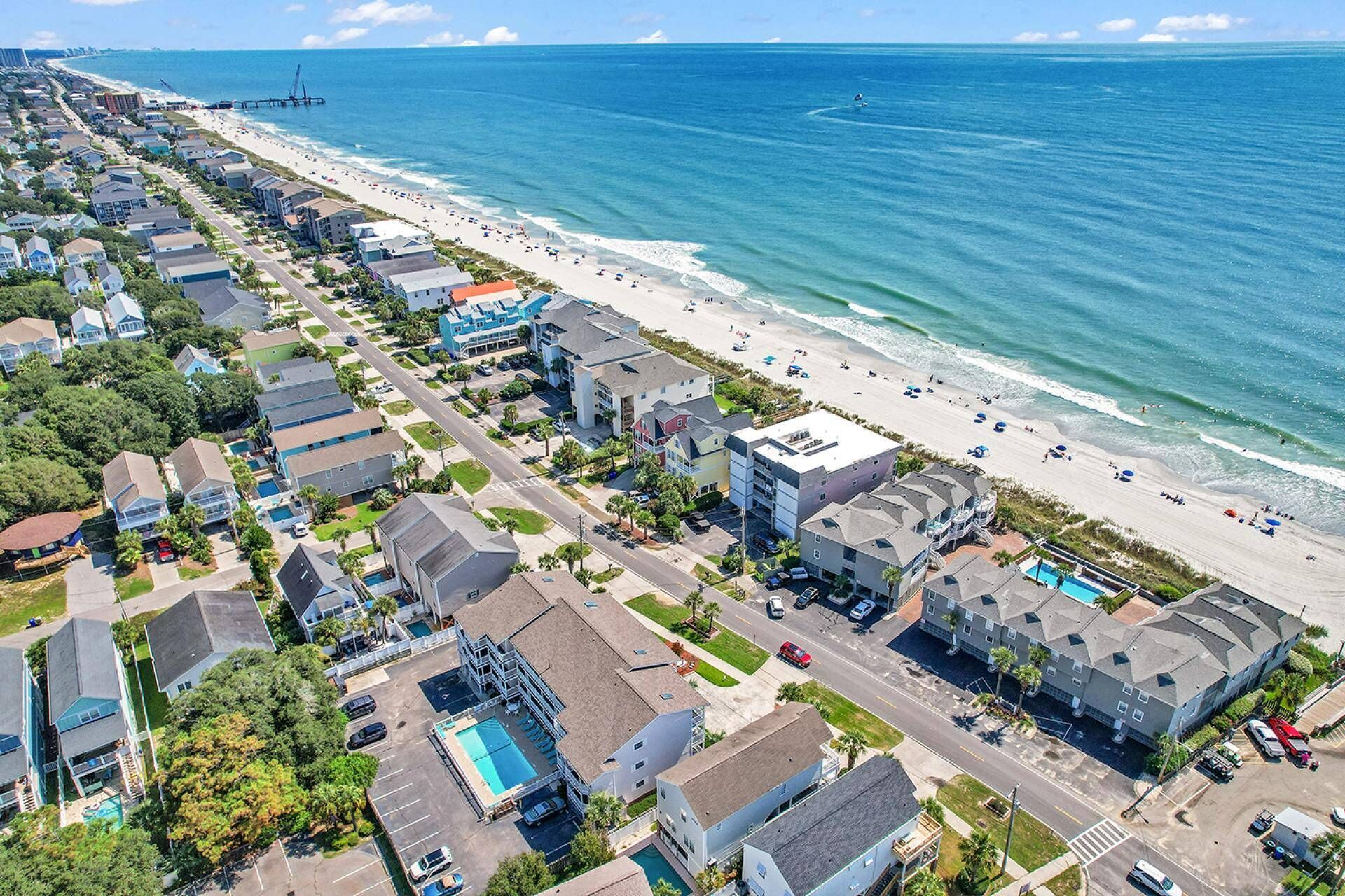 An aerial view of a coastal residential area with a beachfront, including houses, a road, and a swimming pool.
