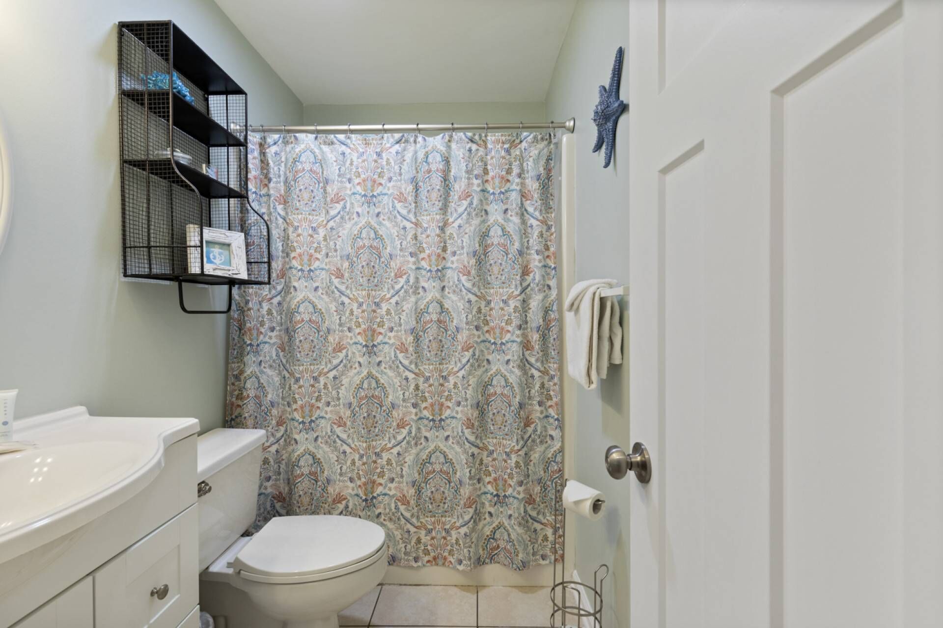 Une salle de bain équipée de toilettes, d’un lavabo, d’un rideau de douche, d’un porte-serviettes et d’une étagère en métal où sont rangés les articles de toilette.