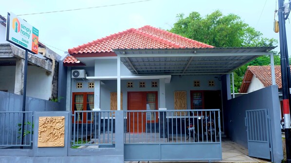 Une maison à deux étages, avec un toit recouvert de tuiles rouges, une véranda et une entrée fermée par un portail.