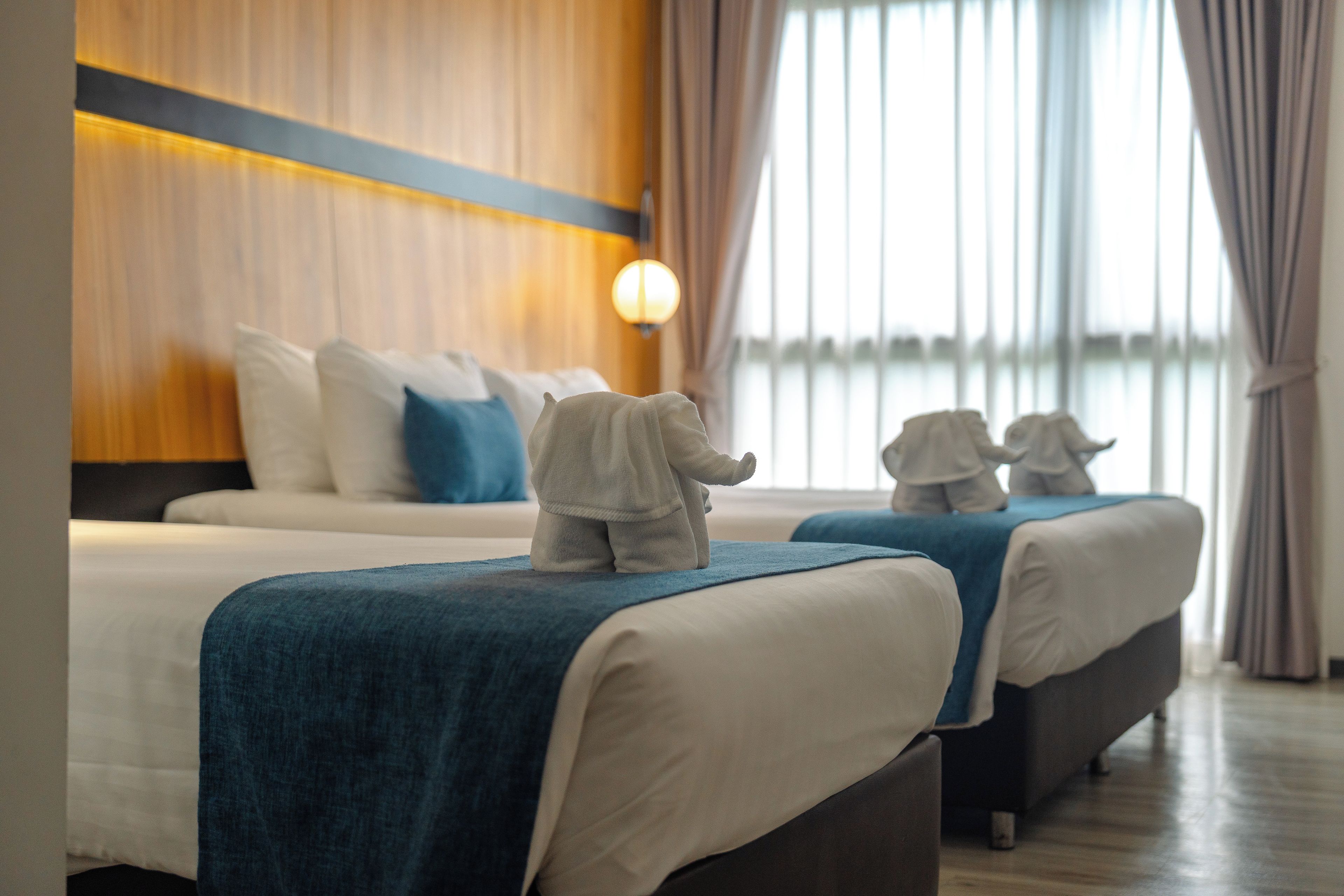 Habitación de hotel con dos camas, una mesita de noche, una lámpara y vistas al exterior a través de amplias ventanas.