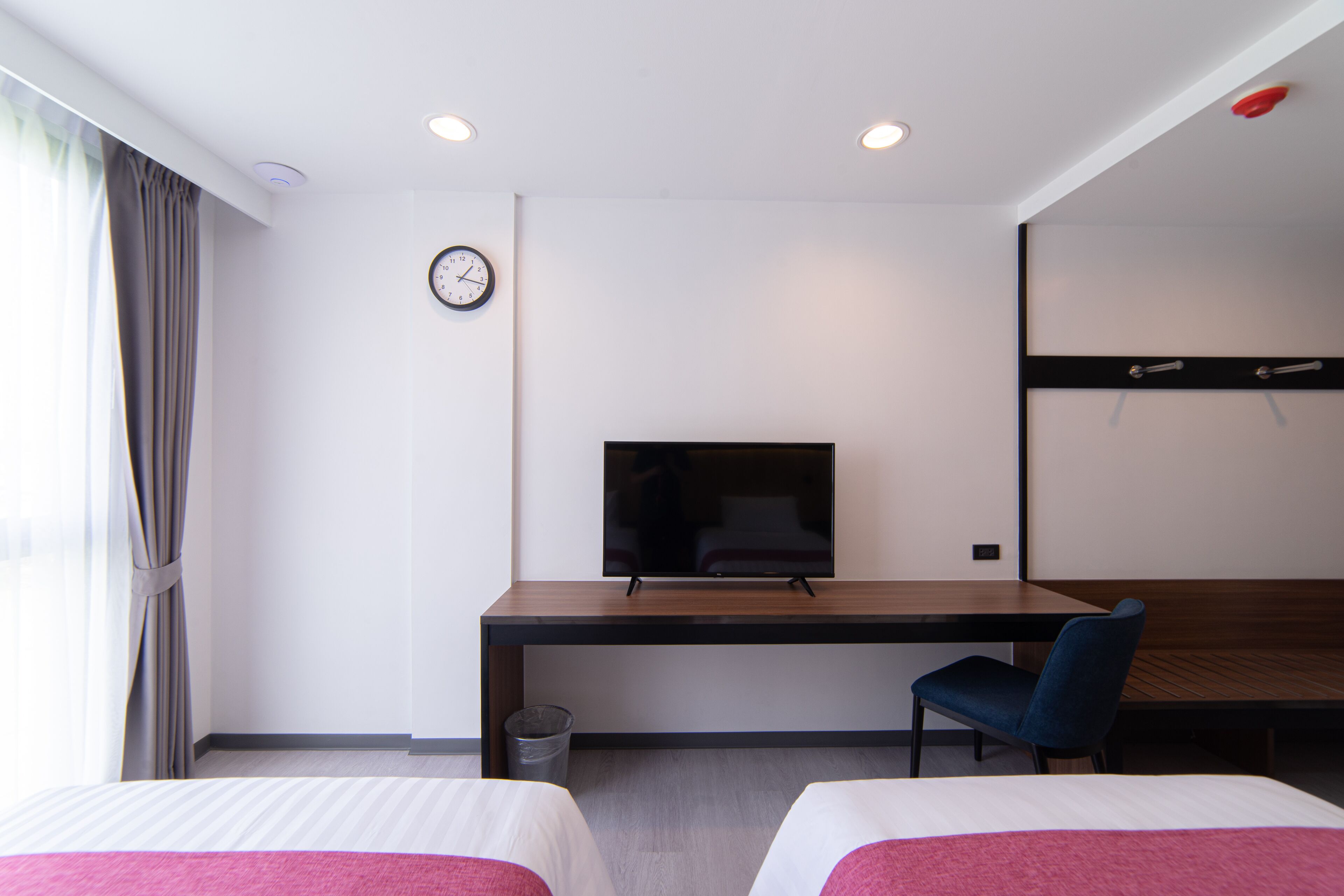 Quarto de hotel com escrivaninha de madeira, televisão, cadeira, duas camas e relógio na parede.