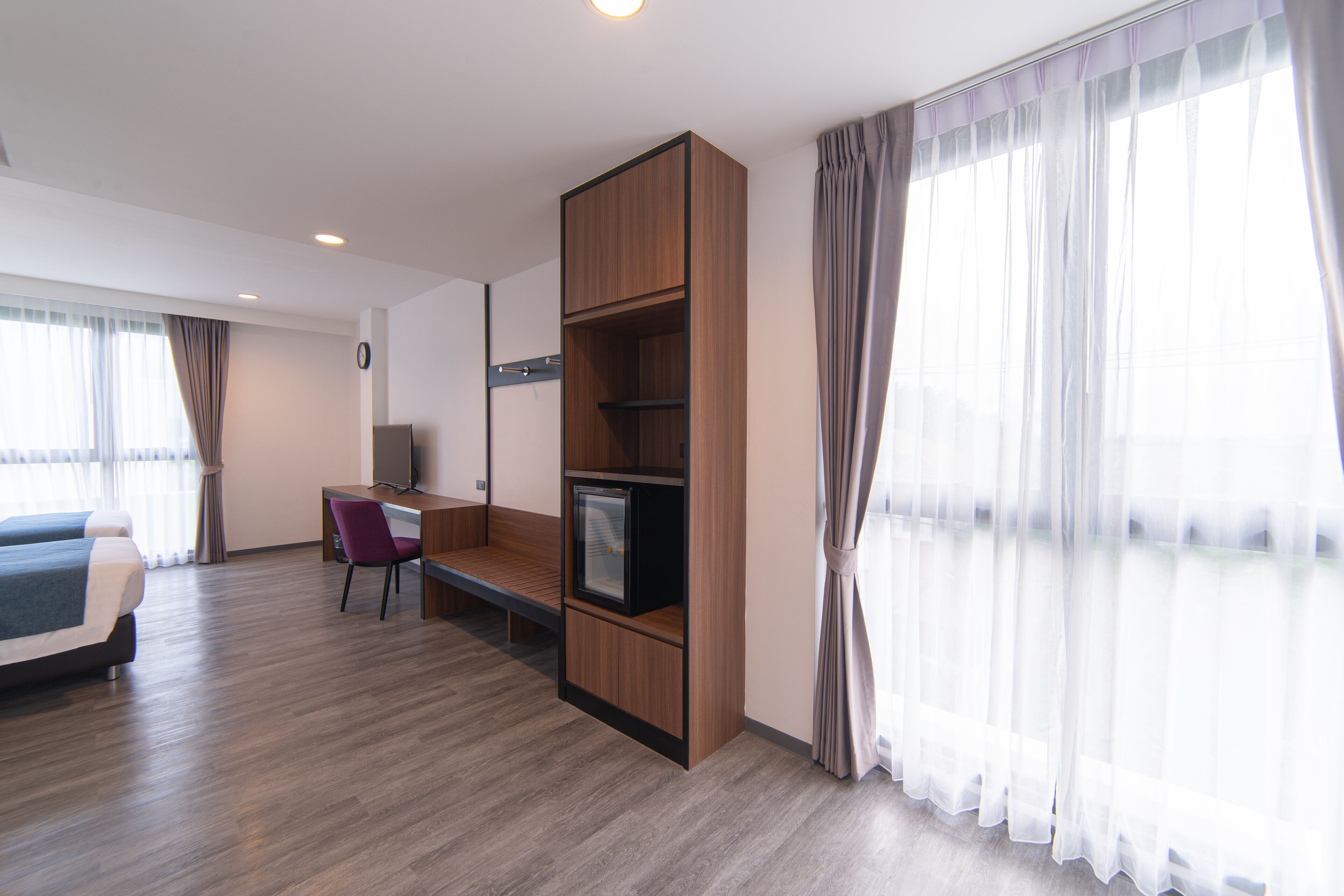 Una habitación de hotel moderna con piso de madera, cama, escritorio con silla, televisor y ventanas amplias con cortinas livianas.