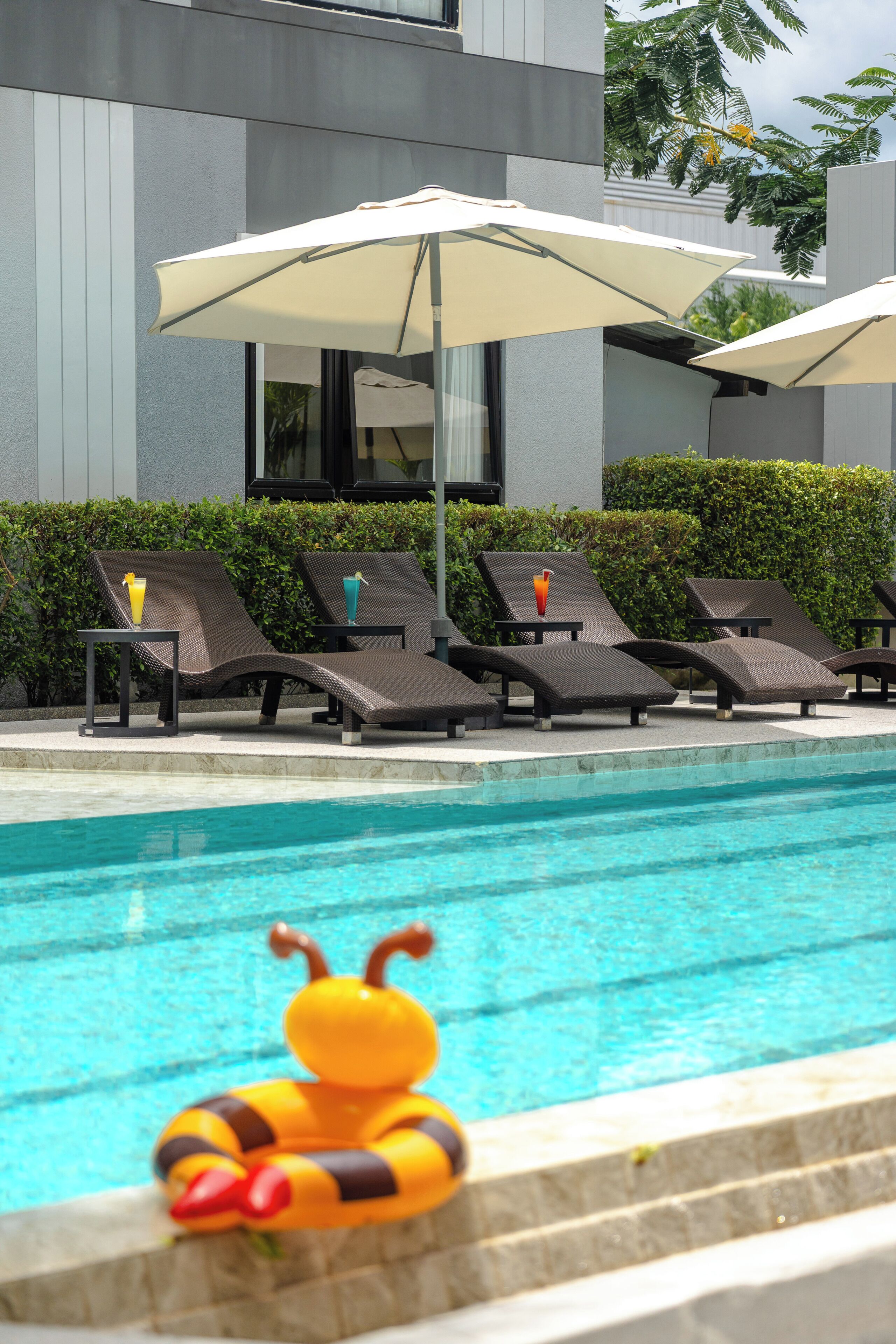 Una piscina con sillas de descanso y sombrillas, un flotador decorativo con forma de abeja y un edificio moderno al fondo.