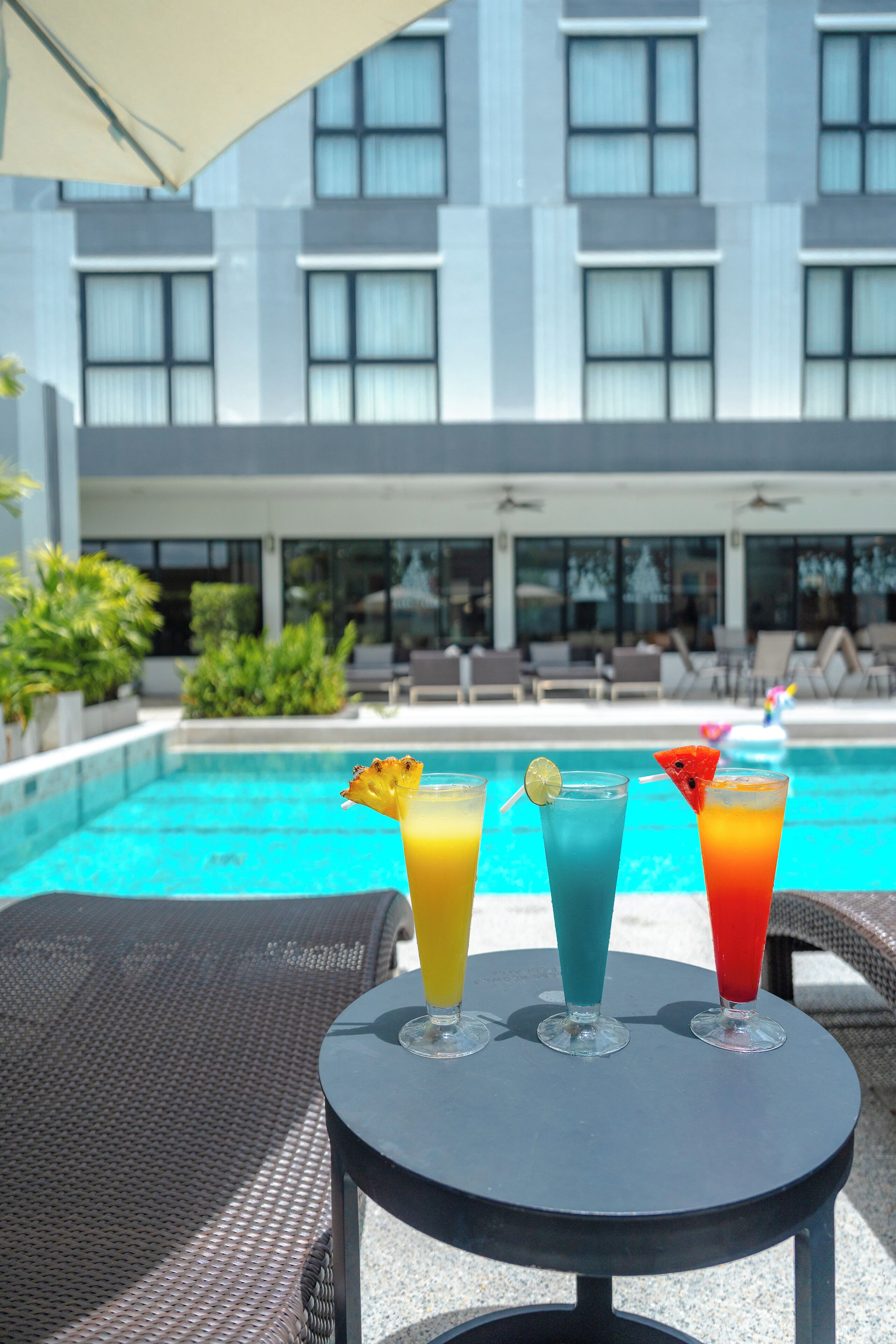 A poolside table with colorful drinks and a modern building in the background.